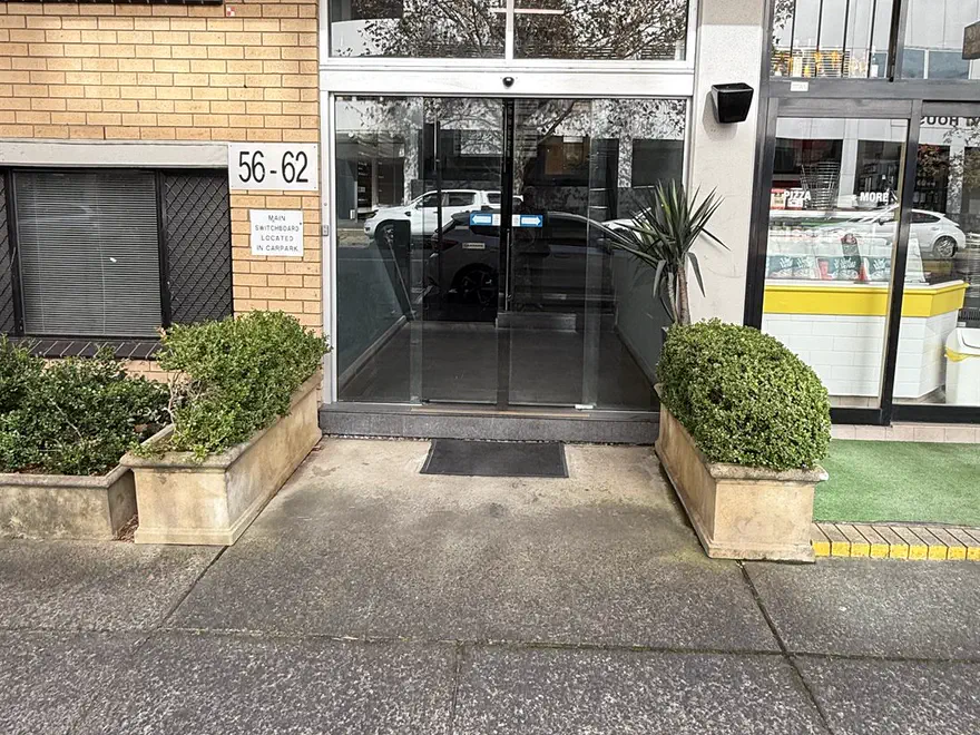 The glass entrance doors beside the number plate 56-62, with a clipped hedge in a planter on each side and a pizza shop front on the right
