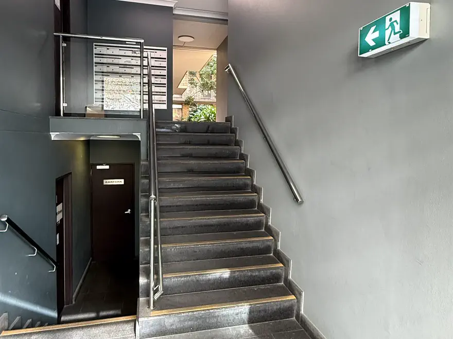 A staircase with a steel handrail rising from the entrance hall, a green exit sign on the right wall and mailboxes at the top